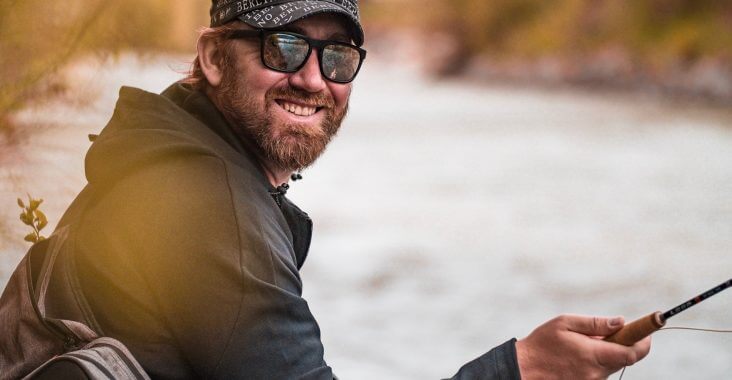 Angler lacht in die Kamera und sitzt an einem Fluss.