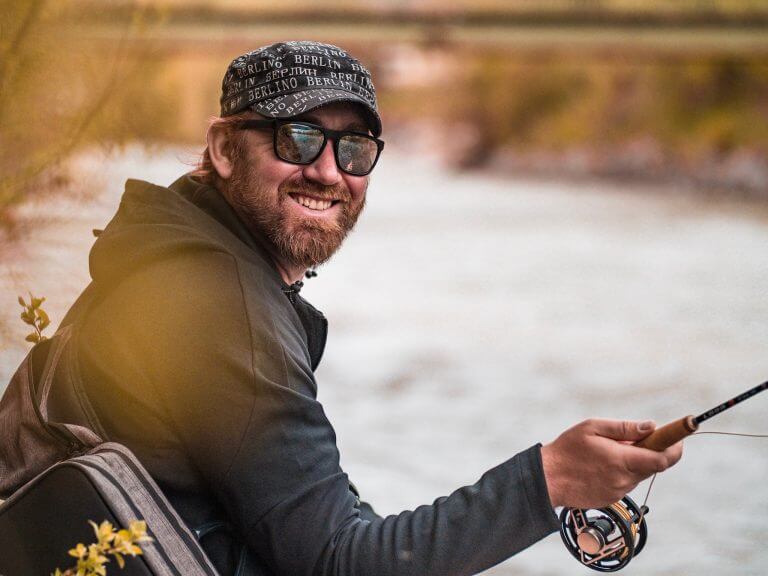 Angler lacht in die Kamera und sitzt an einem Fluss.